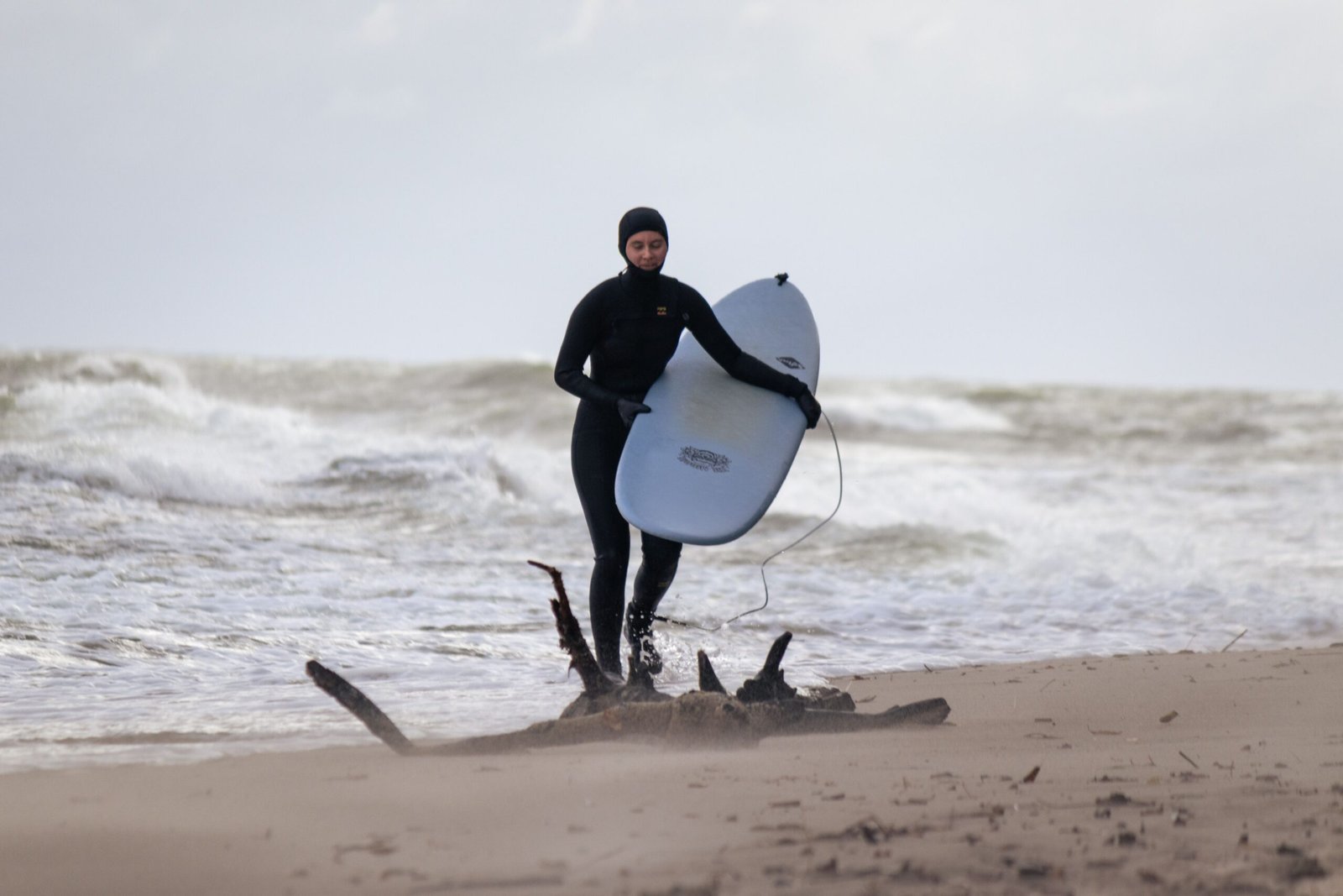 Surfer in a wetsuit walking along a cold, windy beach carrying a surfboard